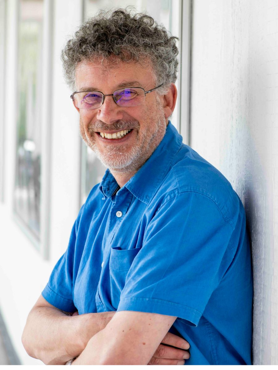 Jeremy Frey portrait profile Jeremy Frey smiling at camera leaning against a white wall, wearing glasses and a blue shirt