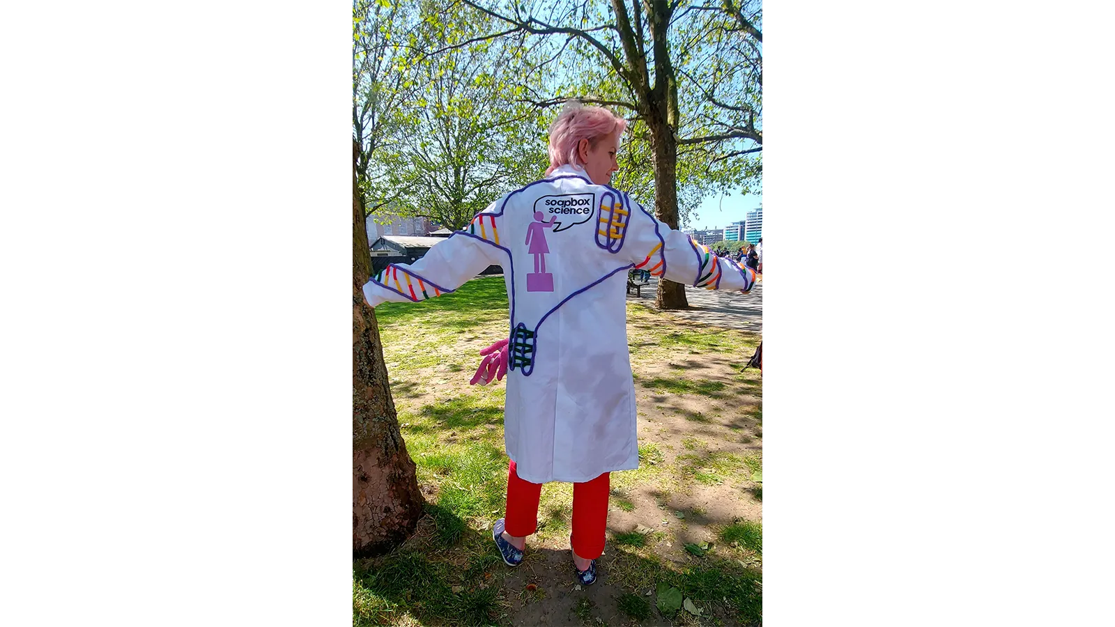 Zoe in a white coat with chemistry markings drawn on it, standing with back to us in sunshine