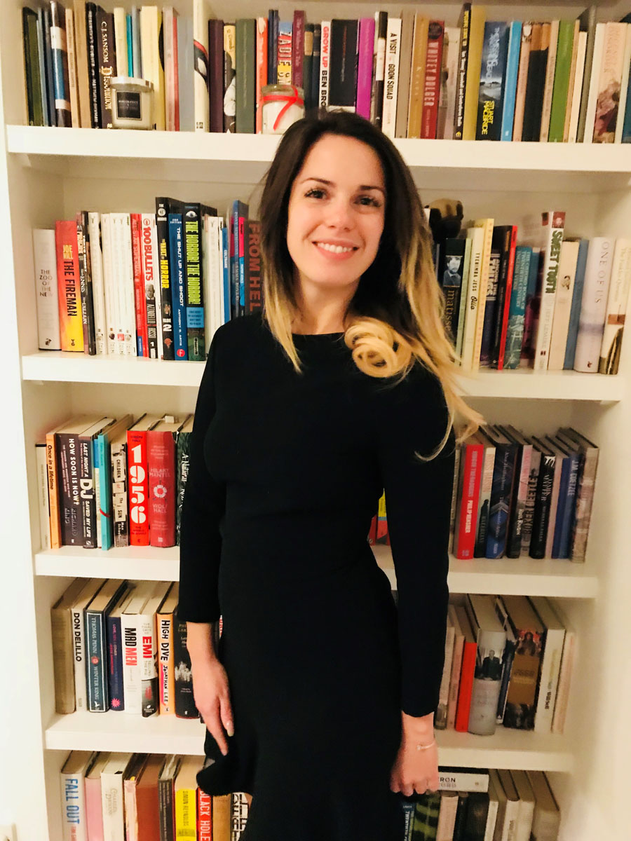 Kristen stands in her home in front of a ceiling-high bookshelf. She is smiling at the camera and wearing a black dress.