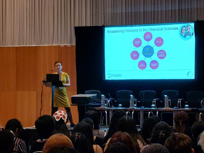 Dr Joanna Jasiewicz gives a speech to the left of a screen showing a graphic with six pink dots and a blue dot Dr Joanna Jasiewicz gives a speech to the left of a screen showing a graphic with six pink dots and a blue dot