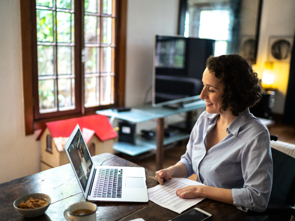 A woman with curly brown hair sits at a rustic wooden table, smiling while looking at her open laptop during a video call.
