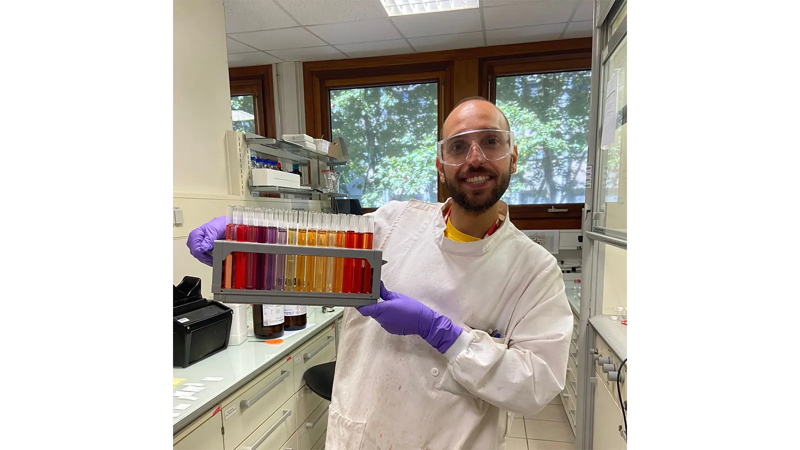 PISCO Team member in a lab coat and gloves holding a tray of test tubes containing coloured liquids
