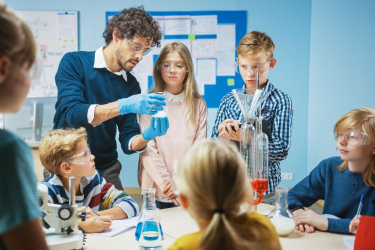 A science teacher teaches five schoolchildren in a classroom, while wearing safety goggles and doing something with glassware