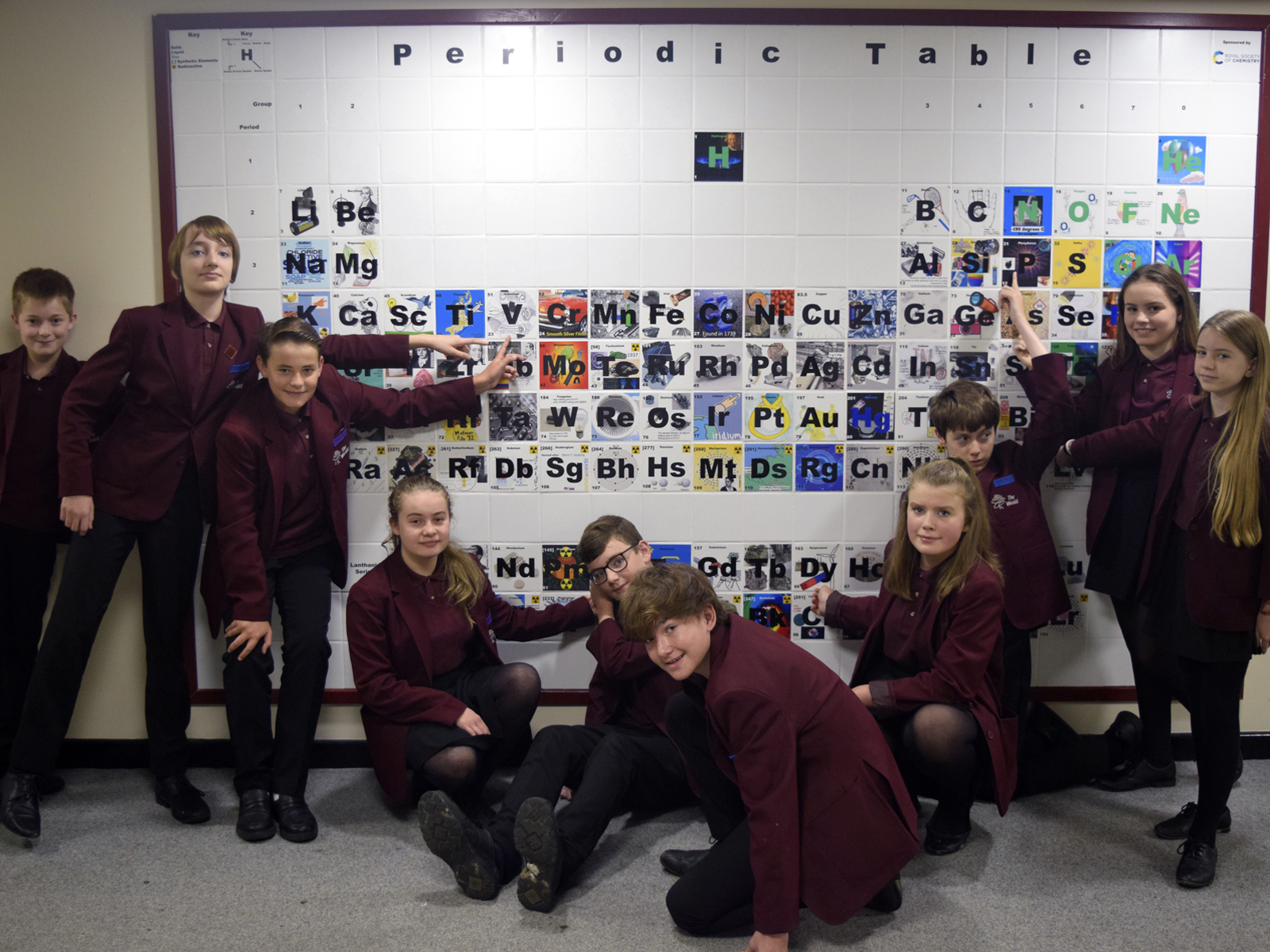 School children in burgundy blazers, some sitting some standing, smile at the camera and point at a large and colourful ceramic periodic table on the wall School children in burgundy blazers, some sitting some standing, smile at the camera and point at a large and colourful ceramic periodic table on the wall