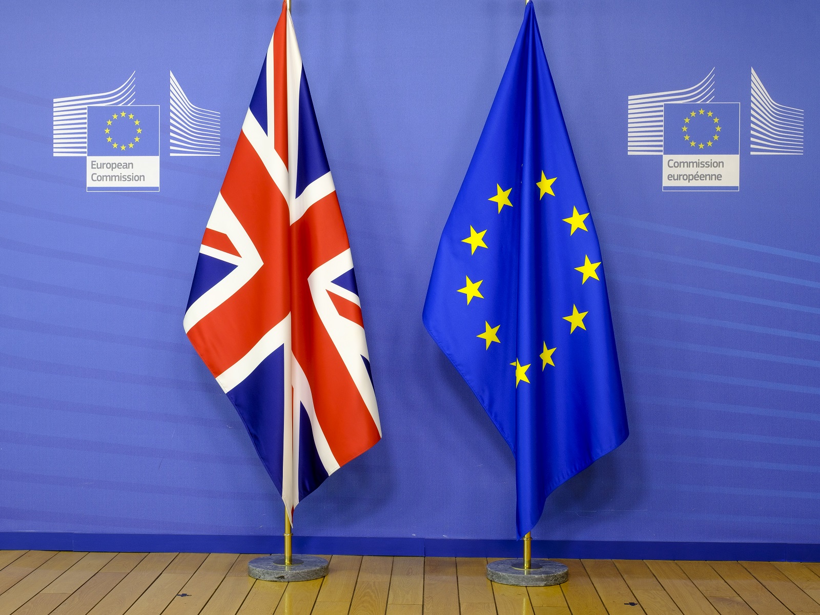 A Union Jack and an EU flag droop from flagpoles in front of a blue European Commission background and above a wooden floor A Union Jack and an EU flag droop from flagpoles in front of a blue European Commission background and above a wooden floor
