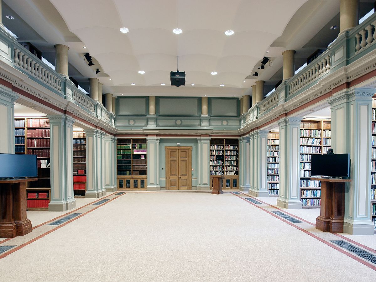 Shelves with books on in library with ornate pillars and cream carpet and balcony
