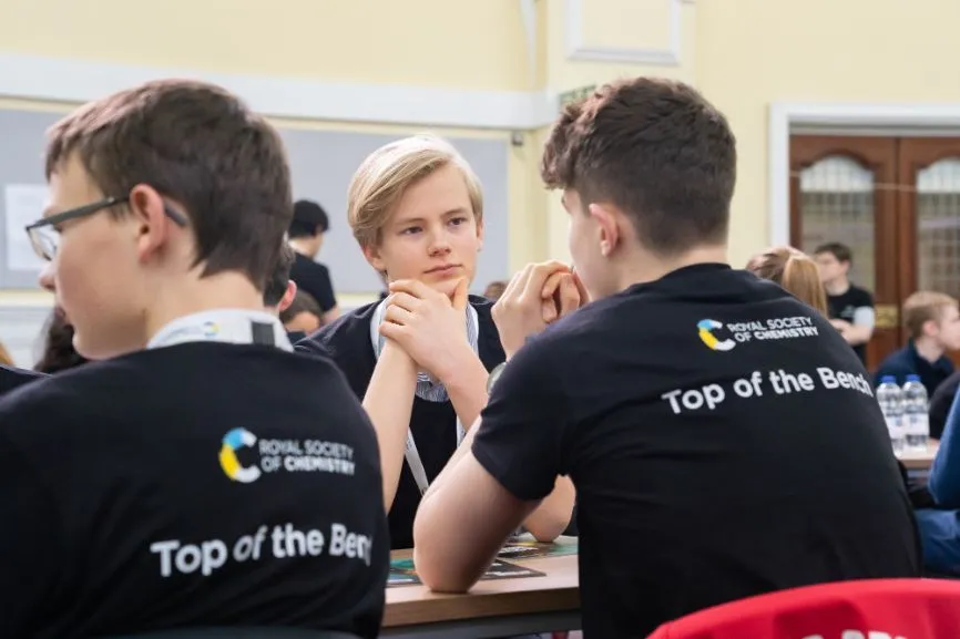 A student thinks about a question while two others, facing away from the camera wear Top of the Bench shirts, during the Top of the Bench final