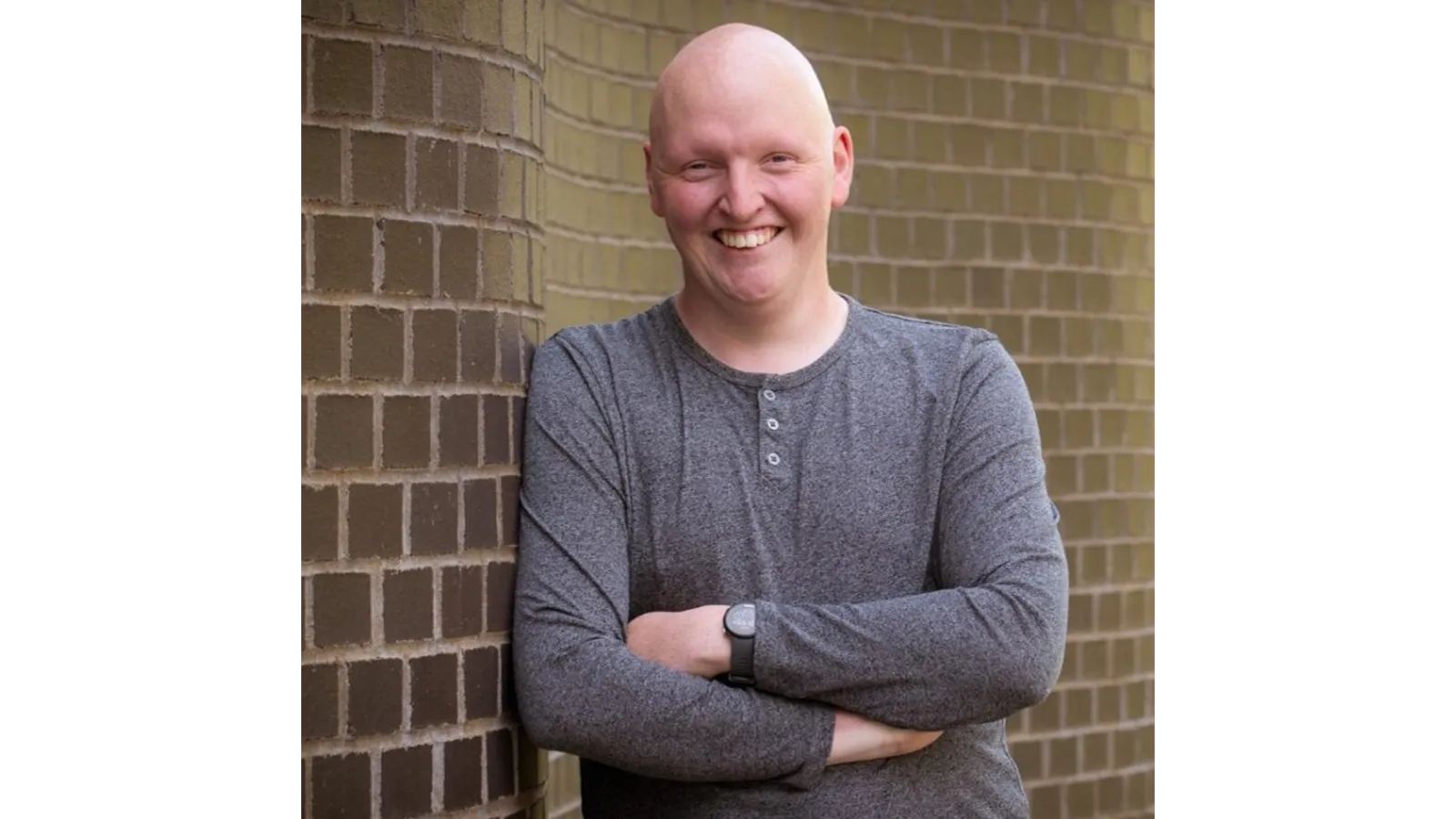 Dr Jack Woolley leaning against a brick wall outsidde, smiling with arms crossed