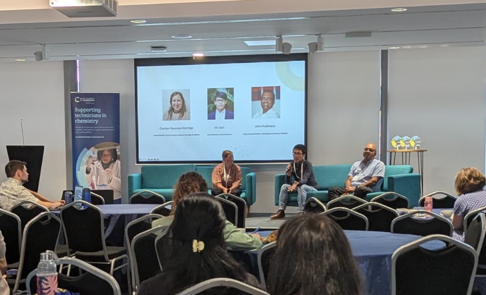The audience sit around circular tables watching a group of three speakers sat at the front of the room taking part in a panel session on AI