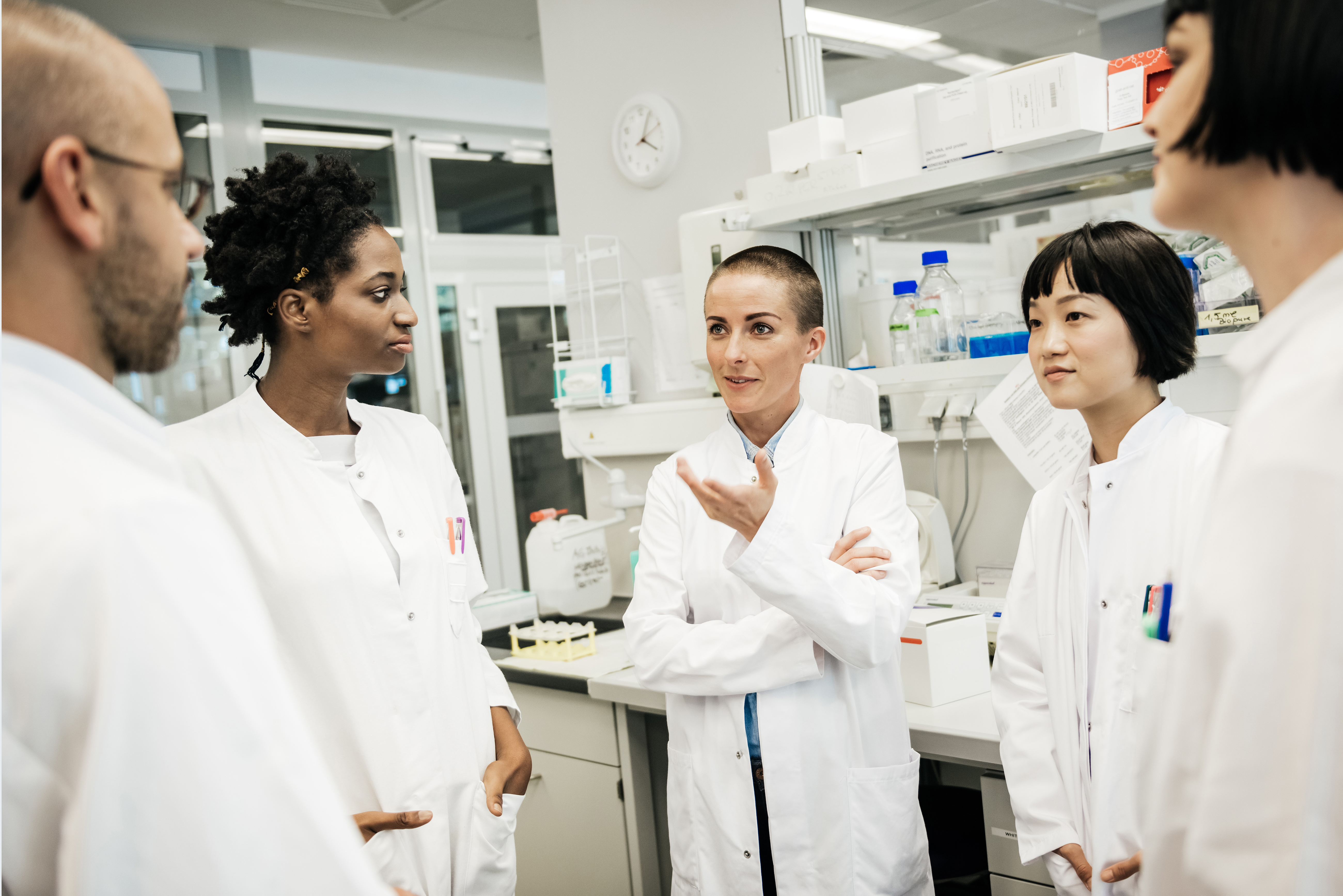 A racially diverse group of five scientists wearing white coats stand and talk in a lab. A south Asian male, a black woman, a white woman, a south-east Asian female and a white woman are visible. A racially diverse group of five scientists wearing white coats stand and talk in a lab. A south Asian male, a black woman, a white woman, a south-east Asian female and a white woman are visible.