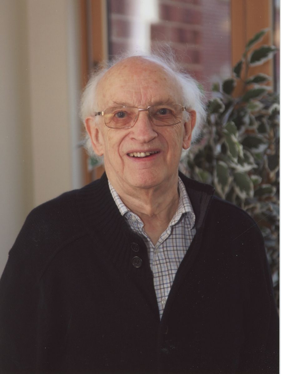 Jack Fennimore standing in front of a plant smiling to camera wearing glasses, shirt and cardigan